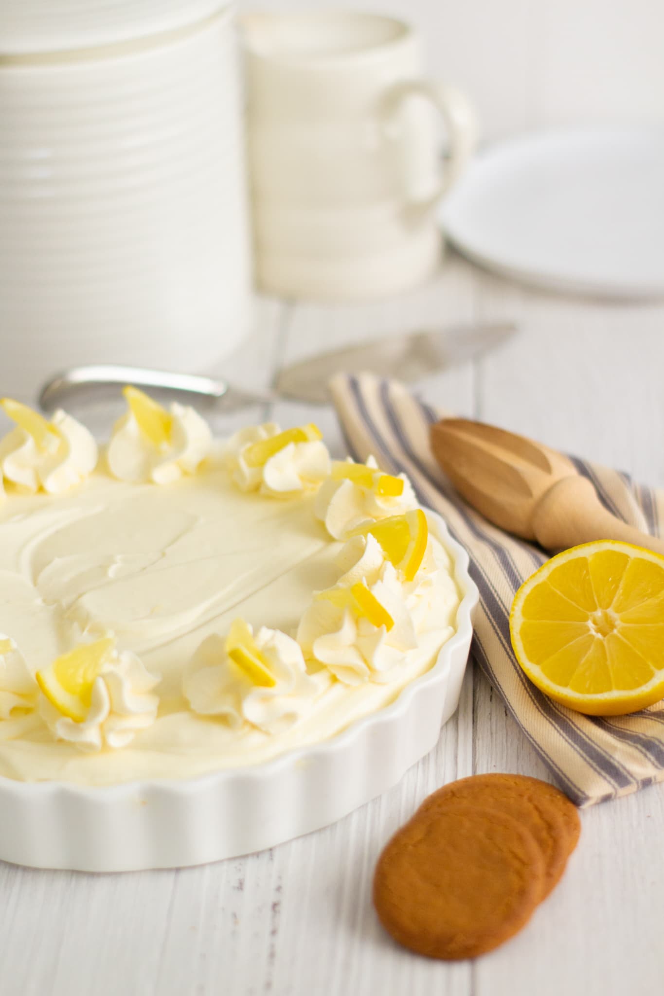 lemon cheesecake in a white dish with ginger nut biscuits in the foreground and a lemon juicer and half a lemon on top of a grey striped napkin