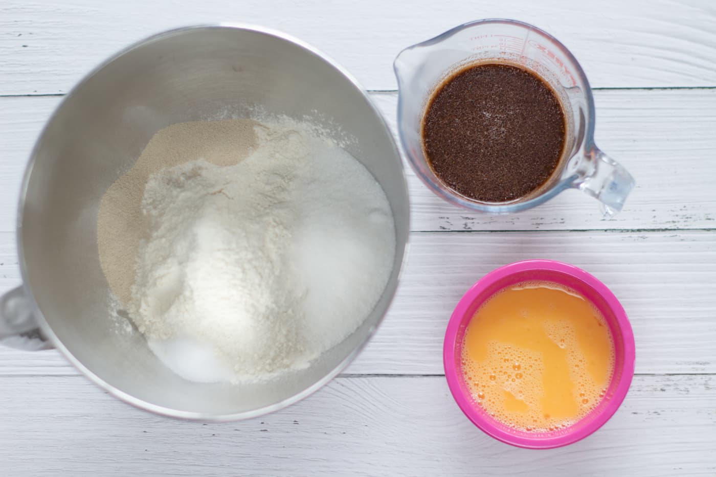 flour, sugar amd yeast in a large metal bowl, dark liquid in a jug containing cinnamon, mixed spice and milk and a pink small bowl with beaten eggs in it.