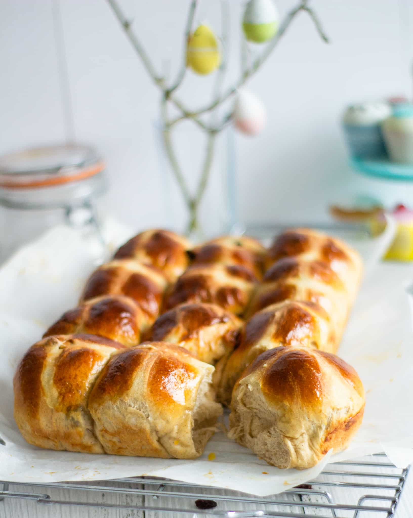 hot cross buns on a wire rack with twigs with small easter eggs hanging on them and cupcakes to the back