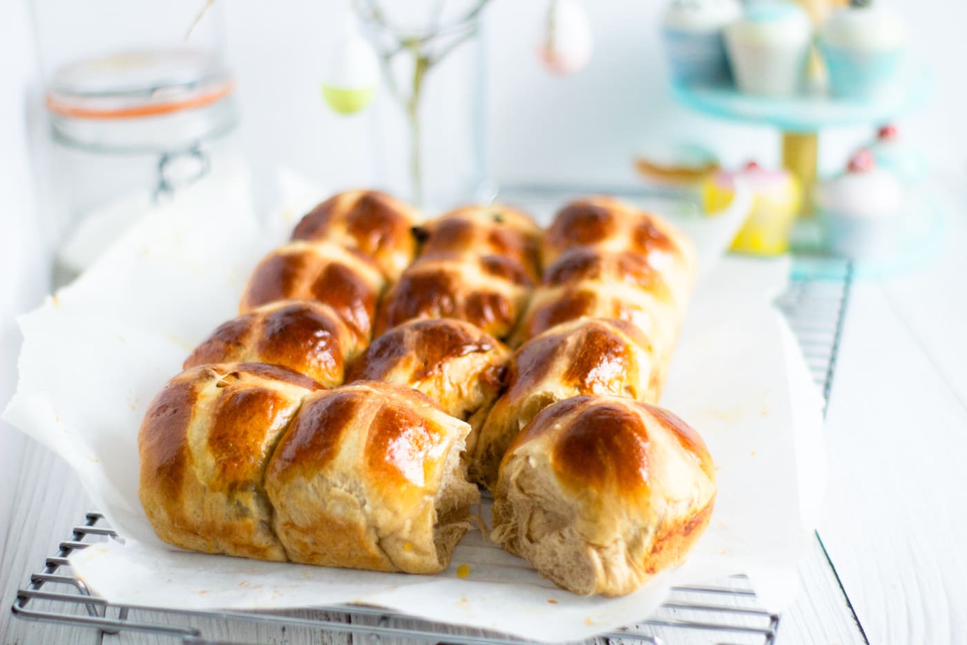 hot cross buns on a wire rack with easter eggs hanging on twigs and cupcakes behind