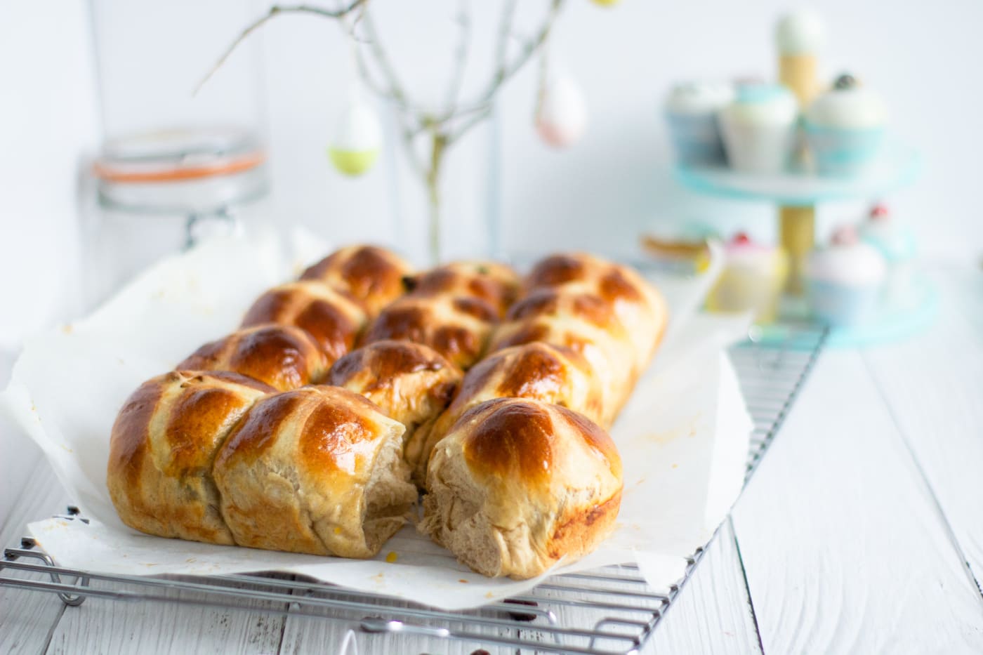 hot cross buns on a wire rack with cupcakes in the backgoround along with a jar of sugar