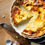 cheese and onion quiche in a white flan dish with a slice cut and a fork and crumbs lying to the side all on a wood surface