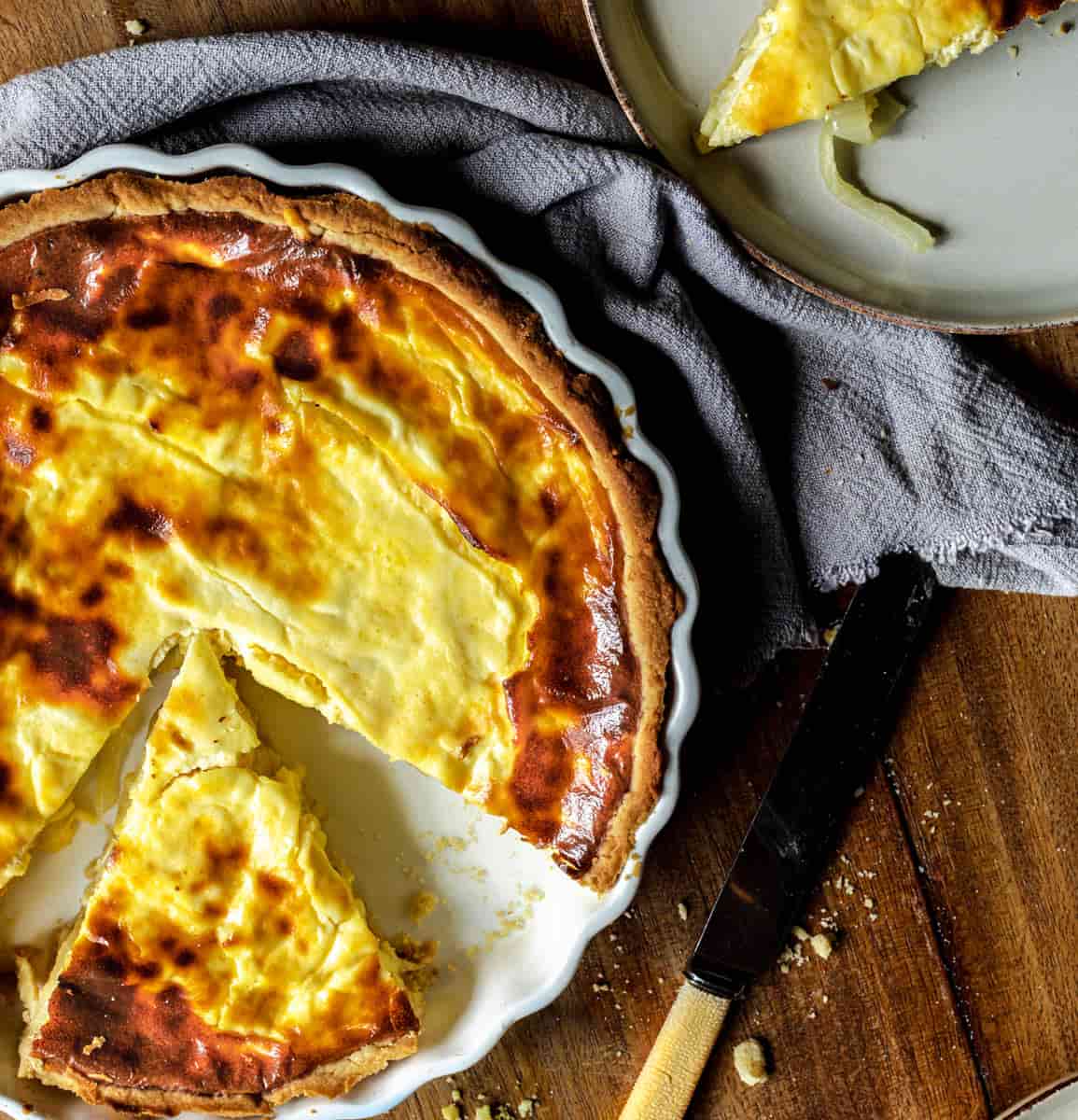 cheese and onion quiche with a slice missing in a white flan dish with a butter knife to the side, a grey napkin is wrapped around with a side plate with a slice of quiche on. Salt is in a small pile on the corner of a small chopping board