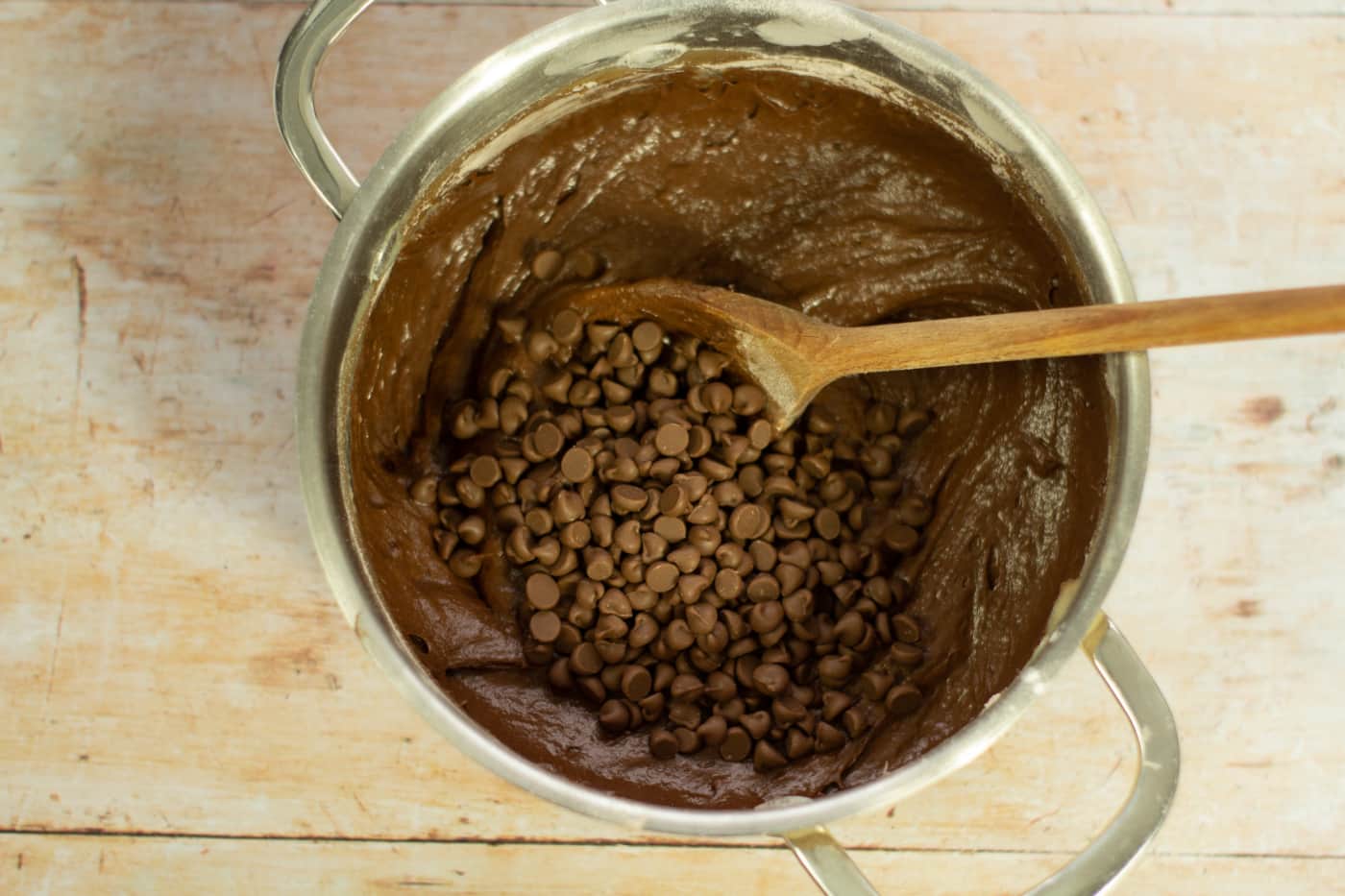 chocolate chips on top of chocolate cake batter in a saucepan with a wooden spoon