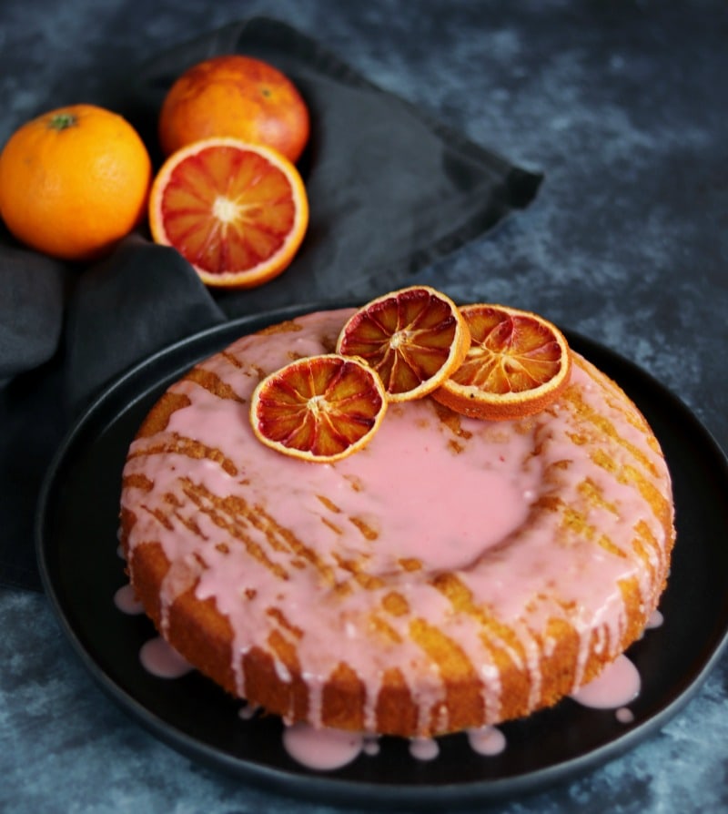 Blood Orange Drizzle Cake on a plate with some oranges on a dark napkin