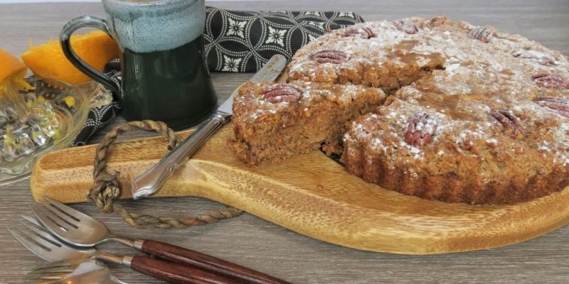 Roast Orange and Pecan Cake on a wooden board with a slice half pulled out and a mug and cutlery near by.