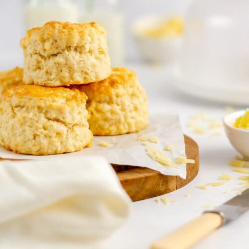 cheese scones on a wooden board with a butter knife to the side and a glass dome behind small dishes of butter are off to the side and behind