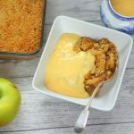 apple crumble with custard in a bowl with bramley apples and a jug of custard nearby.