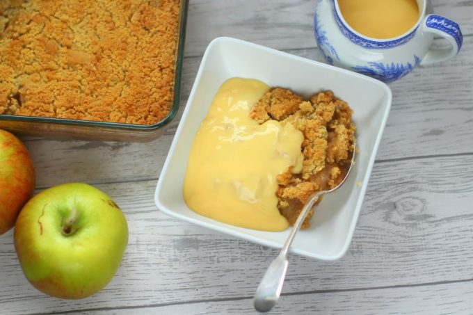 apple crumble with custard in a bowl with bramley apples and a jug of custard nearby.