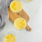 lemon posset in glass bowls on a wooden chopping board with lemon zest scatterd about and a grey napkin swirled at the top of the picture