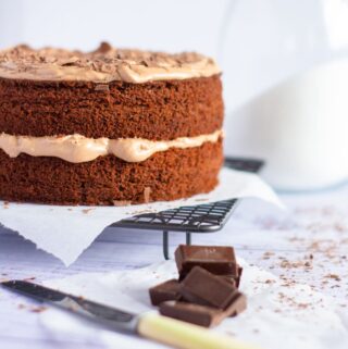 chocolate cake on a wire rack with chocolate chunks and a knife in the foreground and a large sugar pot in the background