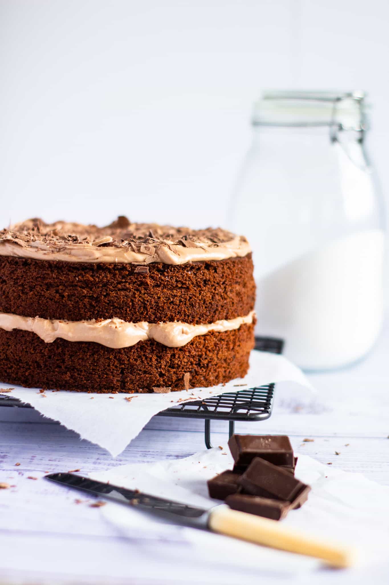 chocolate cake on a wire rack with a butter knife and chocolate chunks in the foreground and a large sugar jar behind