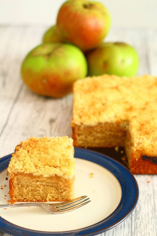 slice of apple crumble cake on a plate with the cake and a pile of apples next to it