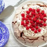 chocolate pavlova topped with cream and raspberries on a serving dish