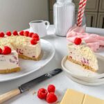 easy no bake white chocolate and raspberry cheesecake on a white plate with a piece missing and placed on a small white plate with white chocolate bar and fresh raspberries in the foreground and white , jar of icing sugar and red and white straws visible in the background jug of cream