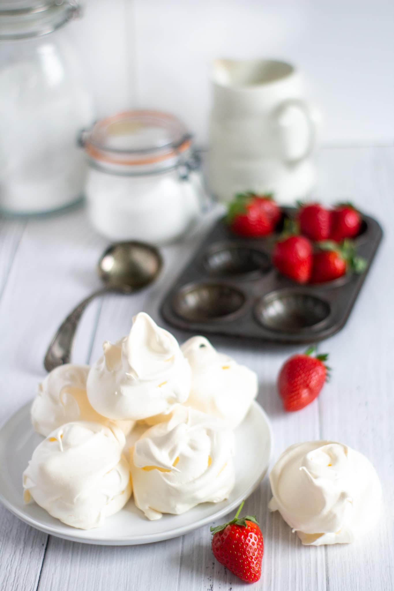 meringues on a plate with strawberries with a metal baking tray behind with sugar pots and a cream jug