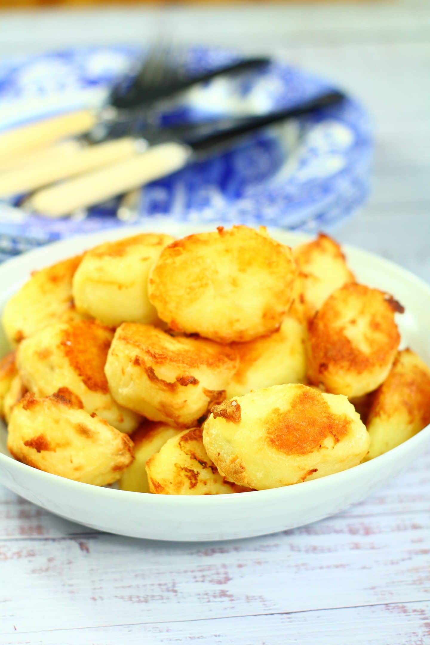 roast potatoes piled high in a white serving dish with plates and knives and forks in the background