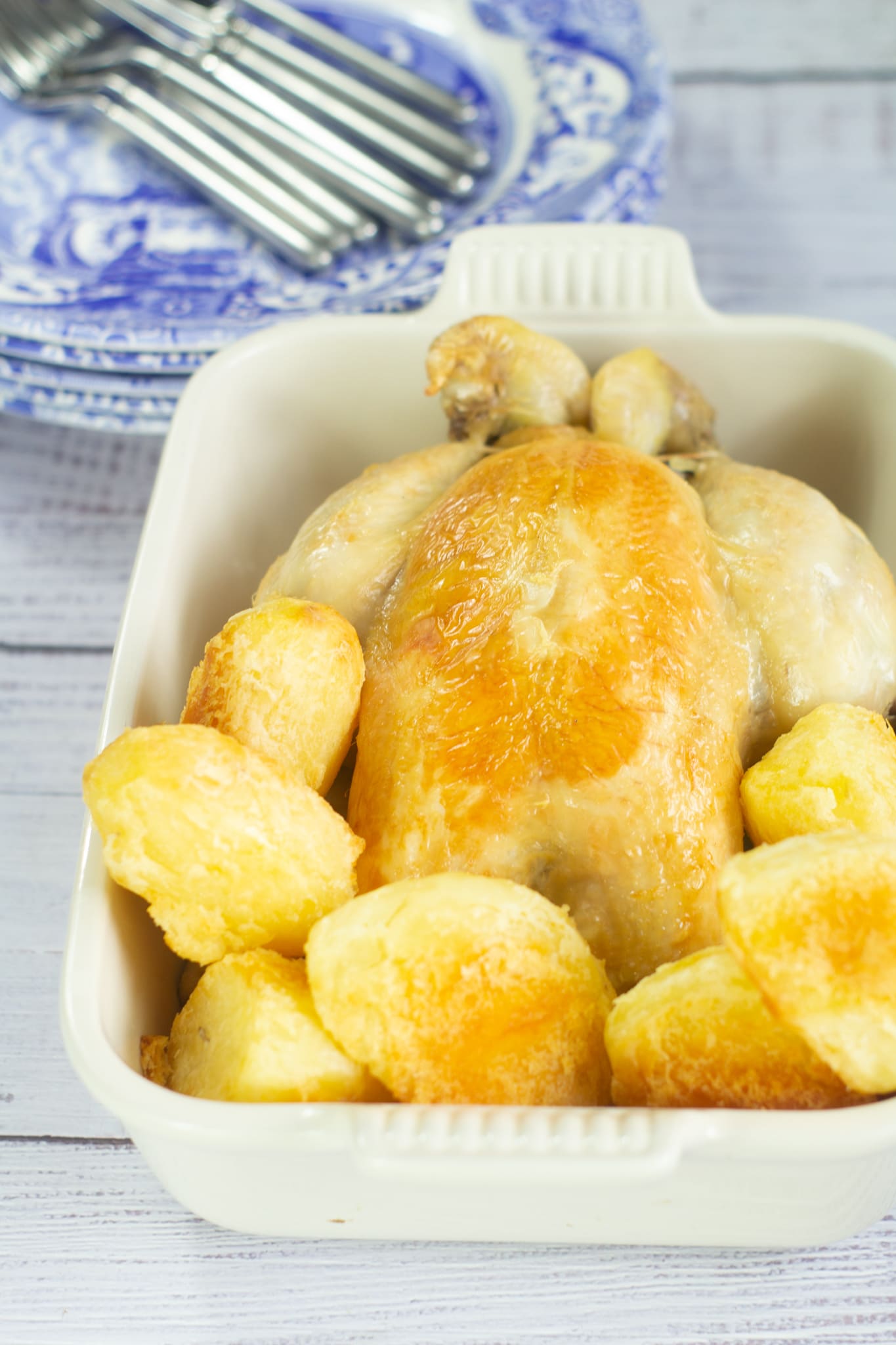 roast chicken in a serving dish with roast potatoes and blue and white plates in the background