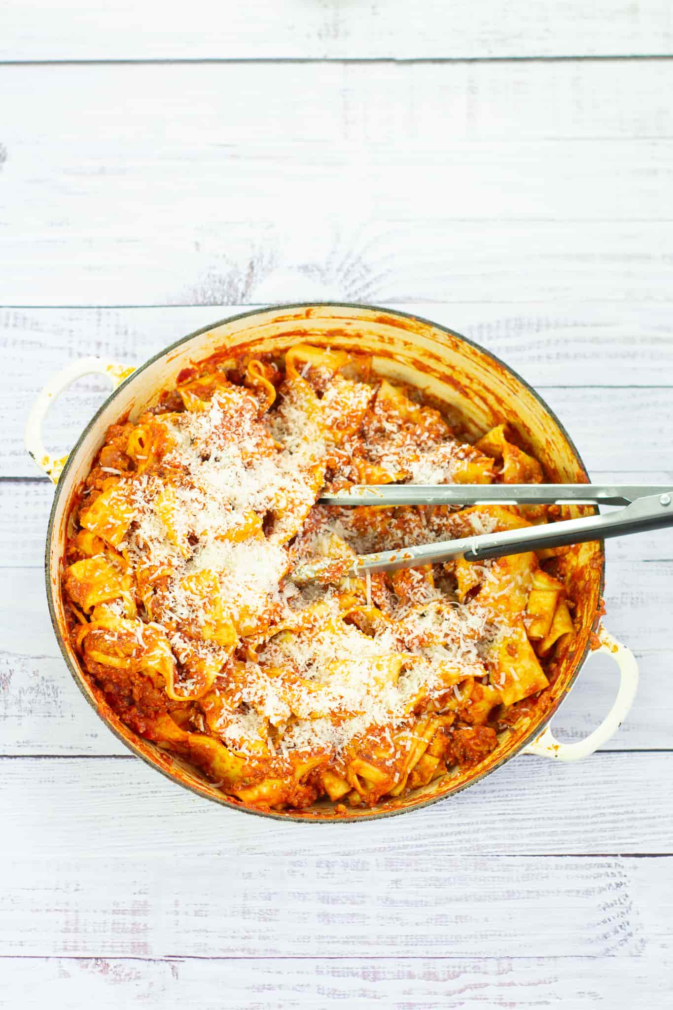 sausage pasta in a pan with tongs and sprinkled with cheese on a white work surface