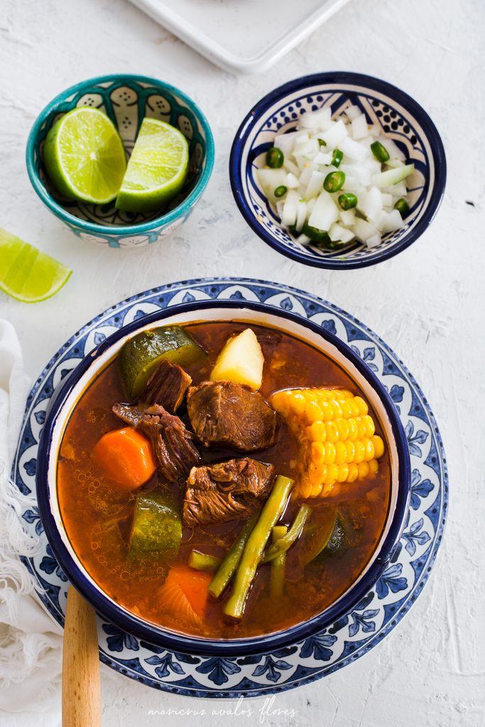 mexican beef soup in a bowl with limes ad onion in bowls above it.