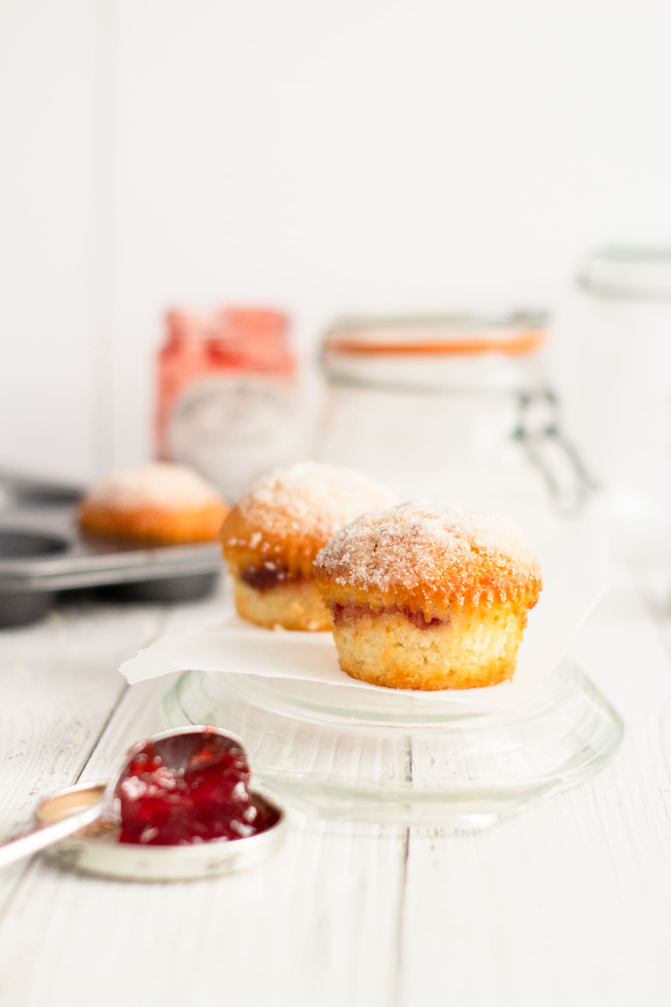 jam duffins on a glass tray with a spoonful of jam in the front and a duffin in a muffin tray and glass jars, jam jar and sugar jar to the back.