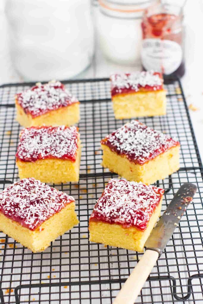 jam and coconut sponge squares on a wire rack with a orange handled butter knife to the side