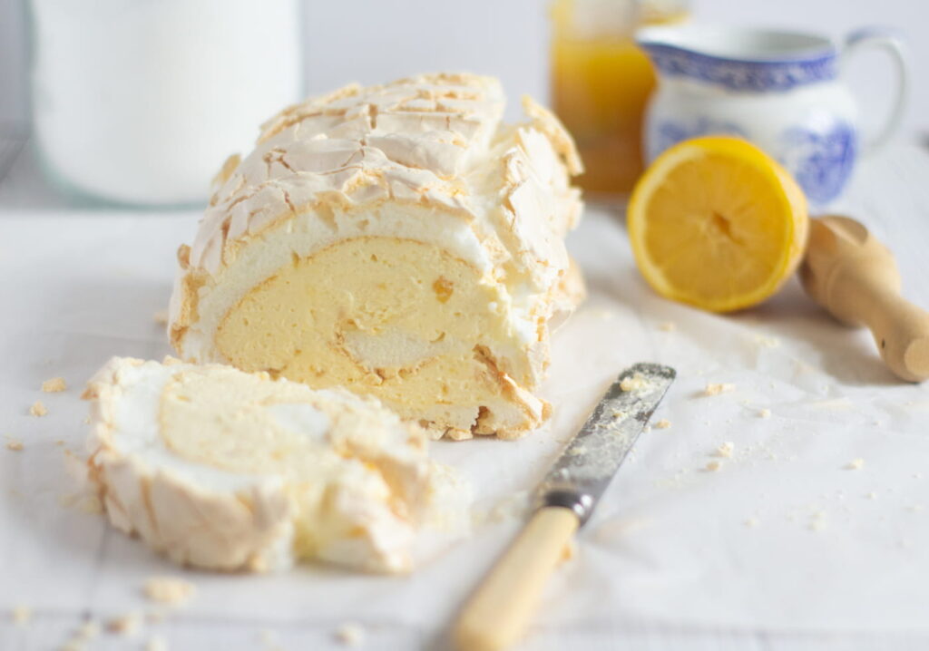 lemon roulade on a white surface with lemons and a lemon juicer in the background and a serving knife in the fore.