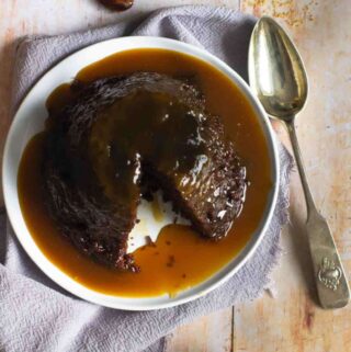 steamed sticky toffee pudding on a white plate on top of a grey napkin and a serving spoon to the side.