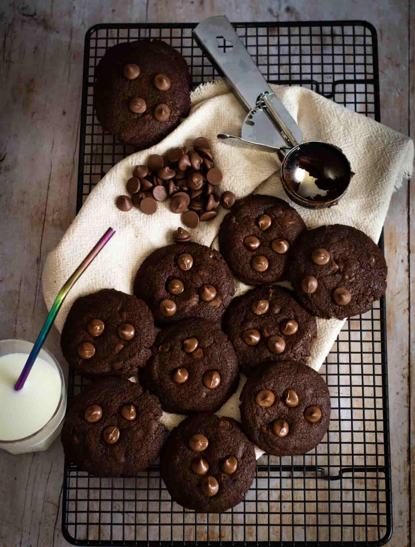 chocolate cookies on a wire rack and cream linen napkin. There is a glass of milk to the left side and an ice cream scoop at the top resting on the edge of the napkin next to some scattered chocolate chips