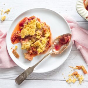 rhubarb and strawberry crumble on a small white plate with serving spoon resting on there all sitting on top of a pink napkin