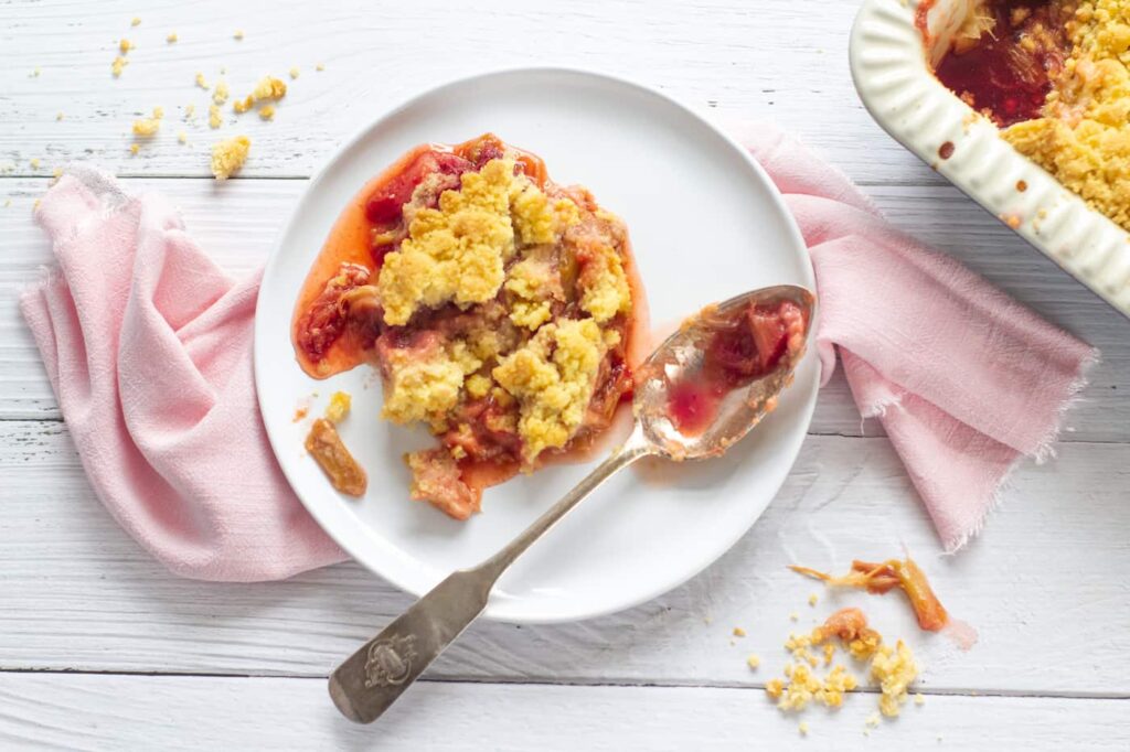 portion of rhubarb and strawberry crumble on a white small plate with a serving spoon all resting on a pink napkin with the corner of the rest of the crumble showing in the top right corner
