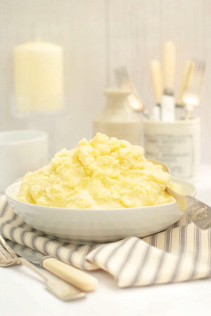 mashed potatoes heaped up in a white serving dish with serving spoon sitting on a grey striped linen napkin. A knife and fork are in the foreground and jars are to the back also containing knives and forks. A glass candlestick and natural coloured candle are to the left.