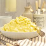 the best mashed potatoes piled high in a white serving dish with a silver serving spoon on the side all on a grey striped napkin. Various jugs and jars behind the bowl and a candle in a glass candlestick to the left.