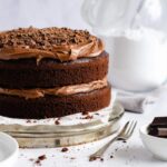 chocolate fudge cake to the left with a cake fork and small white bowl full of dark chocolate chunks to the fore. A jar of icing sugar stands behind.