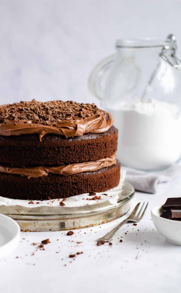 chocolate fudge cake to the left with a glass jar of icing sugar to the right and a cake fork, small white bowl filled with chocolate chunks and cake crumbs in the fore