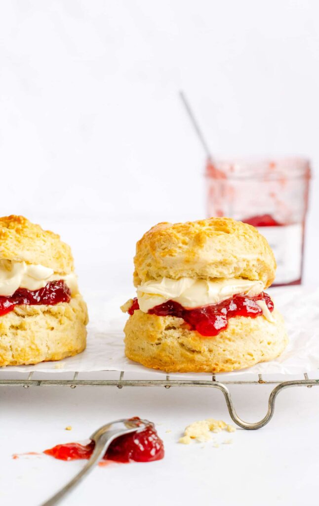 plain scone filled with jam and cream sat on a wire cooling rack with a teaspoon on a blob of strawberry jam in the fore and a jam jar behind
