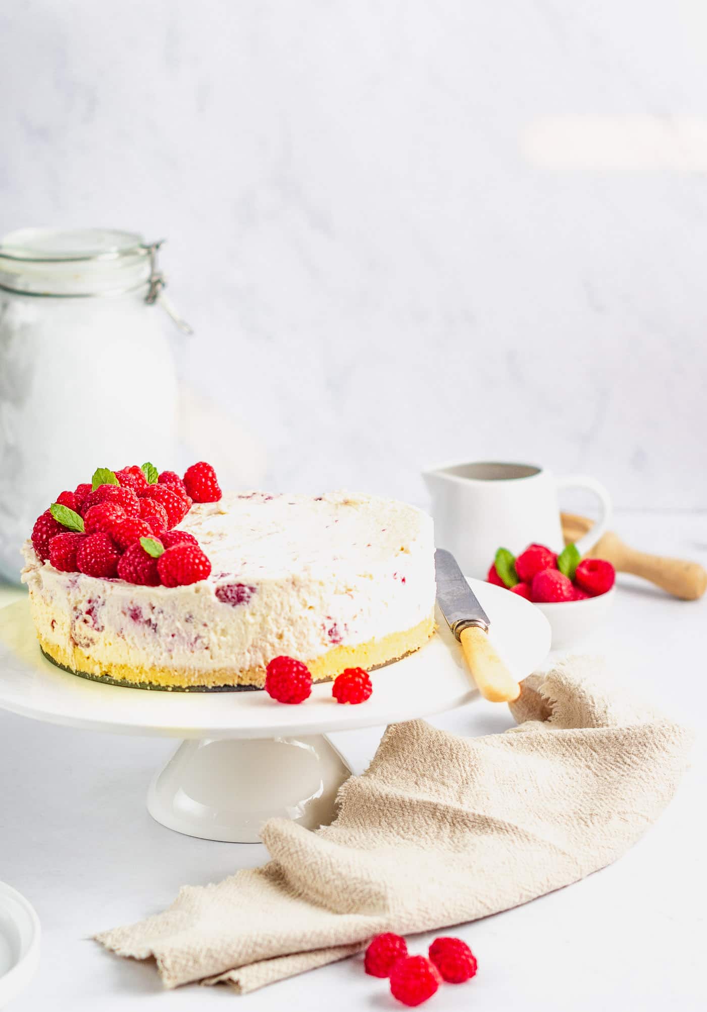 raspberry and white chocolate cheesecake on a white cake stand with a beige napkin t the bottom and a couple of raspberries. A small white bowl of raspberries and a white small jug are behind.