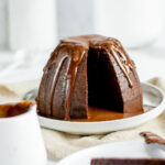 steamed chocolate sponge pudding on a white plate covered in chocolate sauce with a white jug in the foreground and a small white dish with chocolate chunks in it behind.