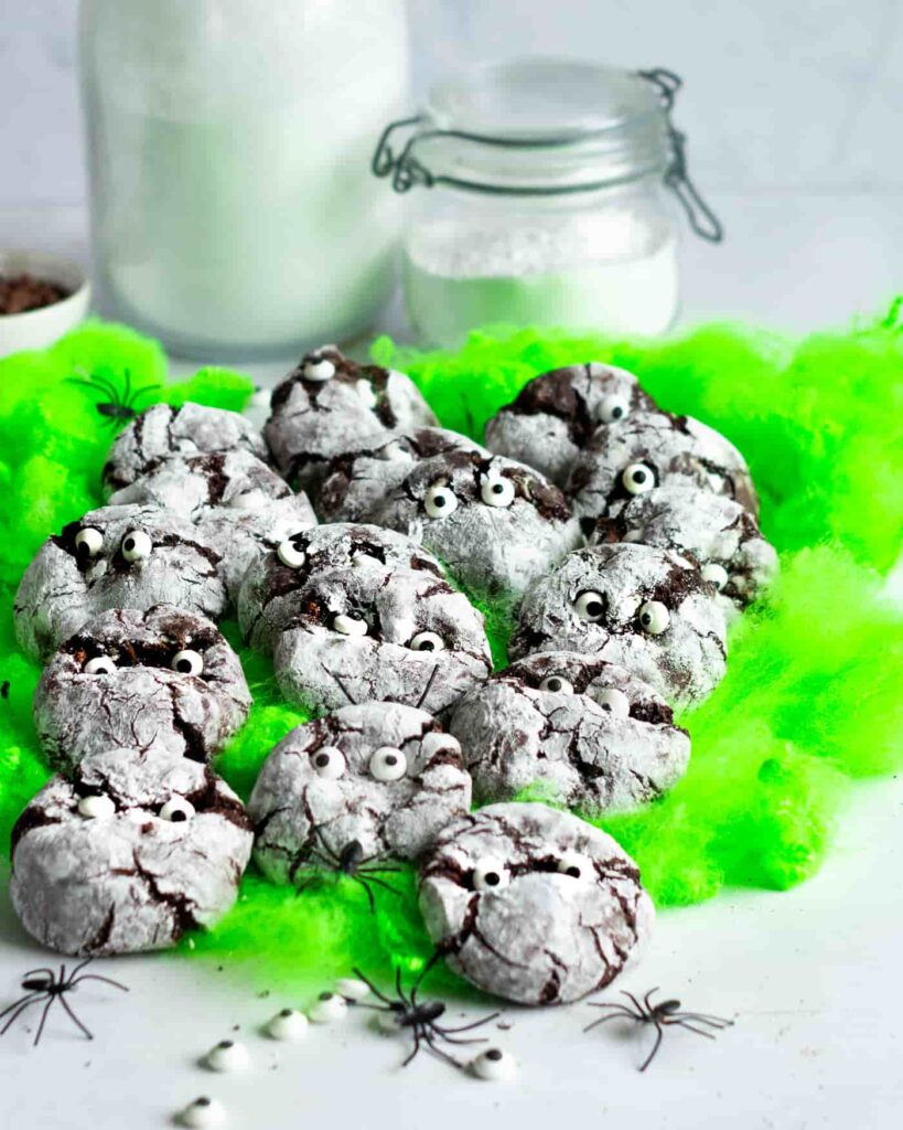 halloween monster mummy cookies laid out on flourescent green cotton wool with spiders crawling over them and two white glass jars in the background.