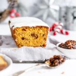 mincemeat loaf cake on a grey napkin with a slice missing from the front of the cake. Silver and red christmas decorations are in the background. A small dish of mincemeat and a spoon are in the foreground.