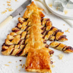 chocolate puff pastry christmas tree on a white surface sprinkled with icing sugar. A knife is to the side with a star cutter on a small white plate above right.