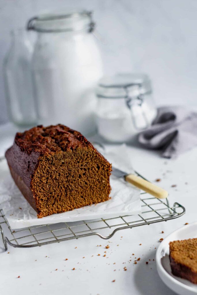 sticky ginger loaf cake on a wire cooling rack with a yellow knife to the side and glass jars with a grey napkin behind, a white plate with a slince of ginger cake is right at the front to the right