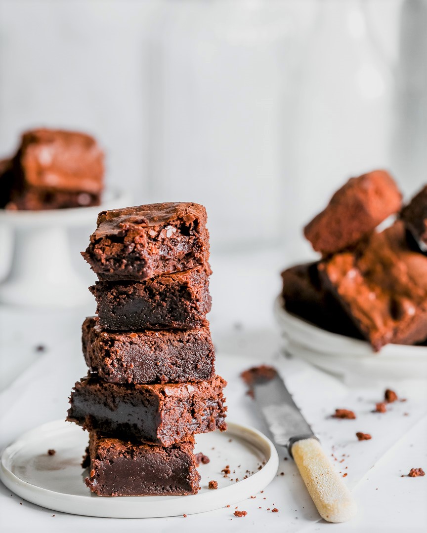fudgy chocolate brownies in a tower on a small white plate with a butter knife to the side and more brownies in the background.
