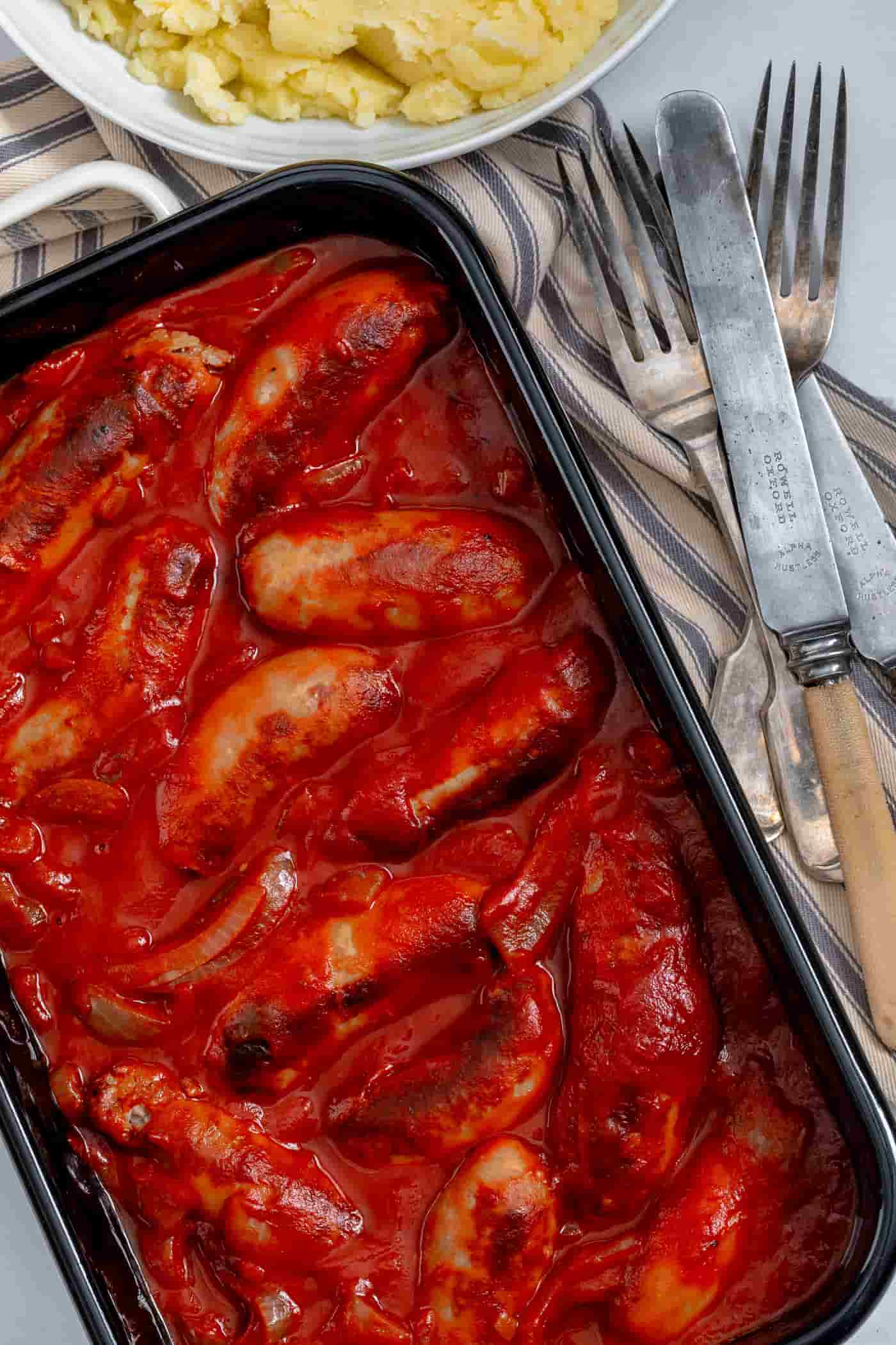 slow cooker sausage casserole in a black enamel baking dish on a striped grey and cream napkin. Knives and forks are to the side with a bowl of mashed potatoes at the top.