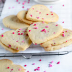 valentine cookies with heart shaped sprinkles scattered over them on a wire cooling rack pile on top of each other