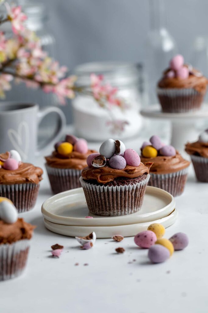 easter cupcakes on top of stack of small white plates with mini eggs scattered around the bottom. Pink flowers are off to the left and a grey mug underneath and more chocolate cupcakes behind.