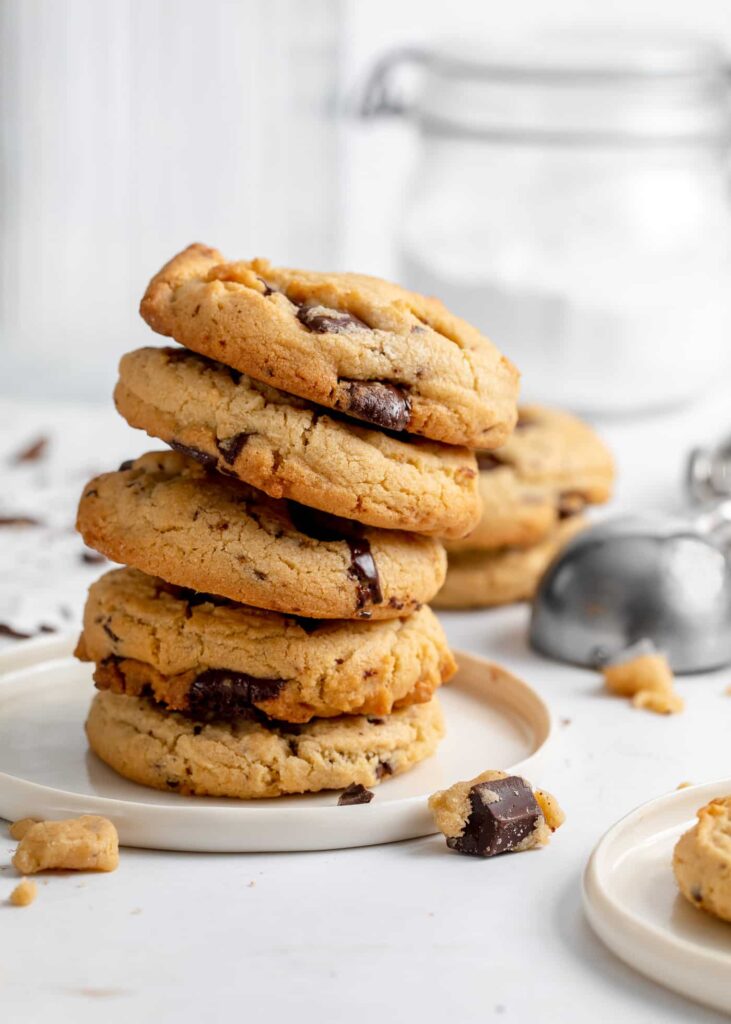 stack of peanut butter cookies with a small lipped white plate with a cookie on the side and a ice cream scoop and glass tall jar in the background