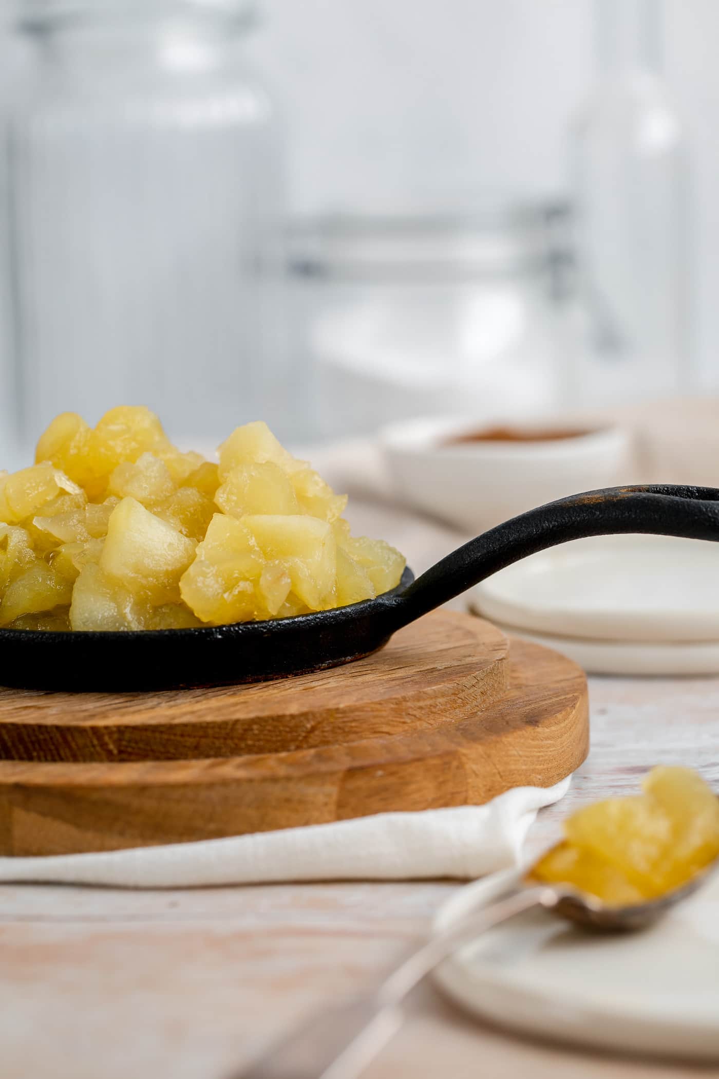 stewed apples on a small black skillet on a round wooden chopping board with glass jars and small white plates in the background. A spoon with stewed apples on lies on a small white plate to the fore.