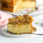 slice of rhubrab crumble cake on a small white lipped plate with another plate and slice of cake partially showing to the right. A pink napkin is visible to the left and the rest of the cake sits on a wire cooling rack on top of baking paper in the background.