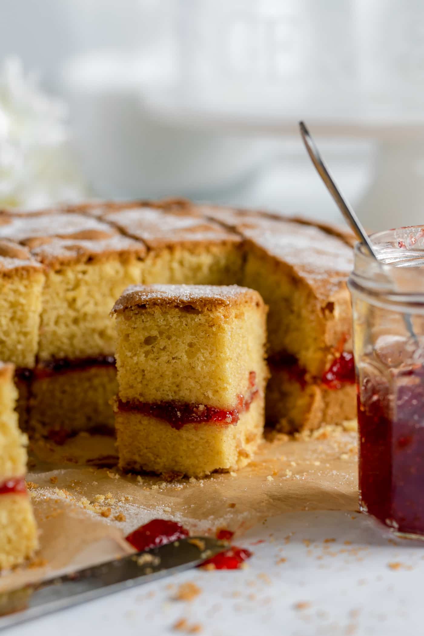 victoria sponge traybake on brown baking paper with a jar of jam with a spoon in it to the right side and a couple of rectangular pieces of cake standing in front of the main cake. A knife with a blob of strawberry jam lies in the front of the picture